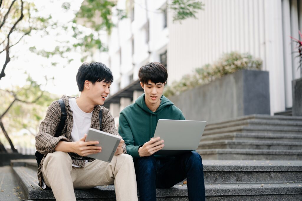 Two smart young Asian college students focusing on his school project, looking at laptop and tablet